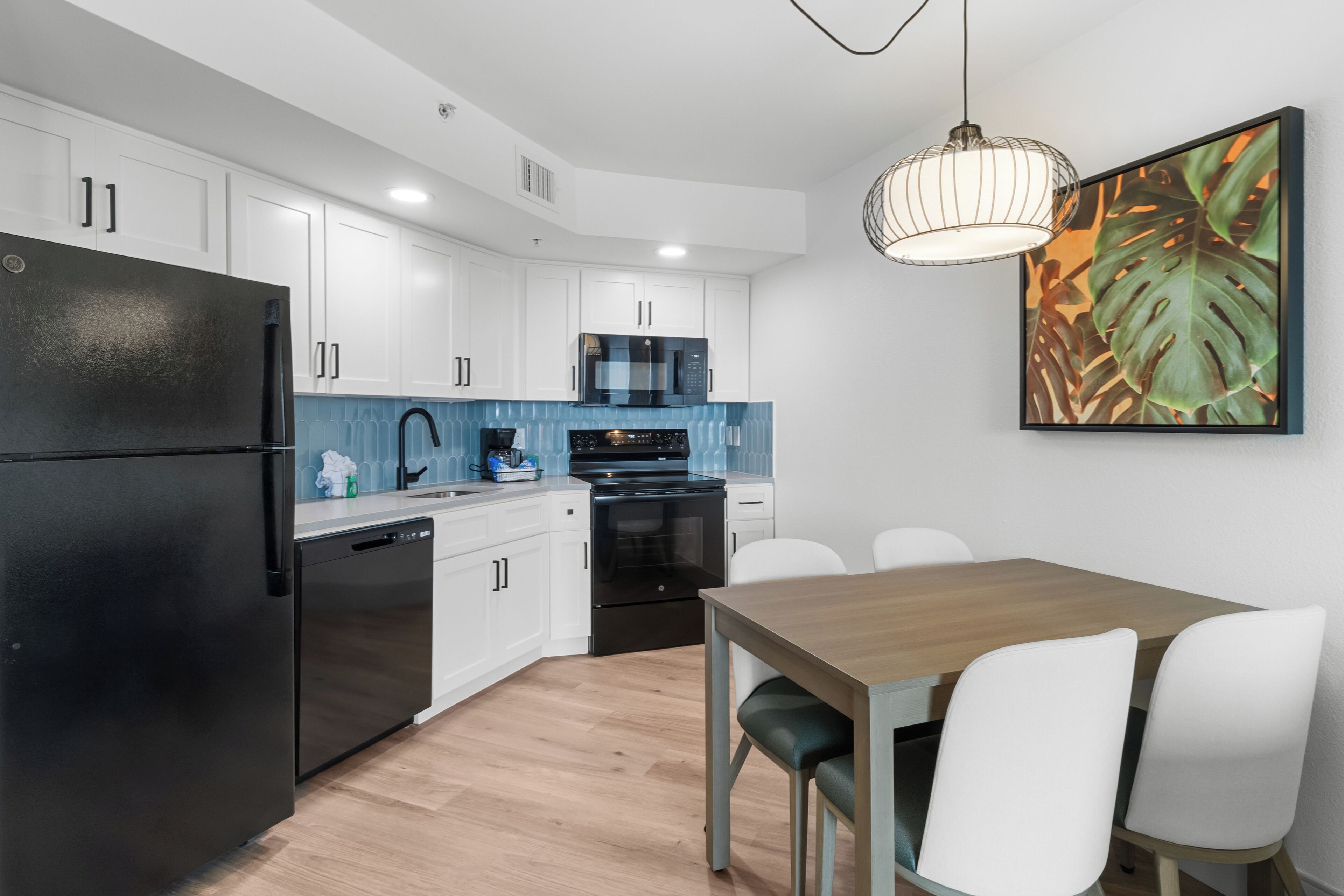 A modern kitchen with white cabinets, a black refrigerator, a microwave, and an oven. There is a dining table with white chairs and a hanging light fixture above it.