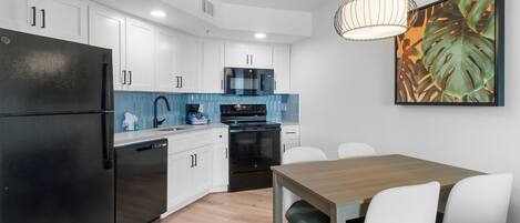 A modern kitchen with white cabinets, a black refrigerator, a microwave, and an oven. There is a dining table with white chairs and a hanging light fixture above it.