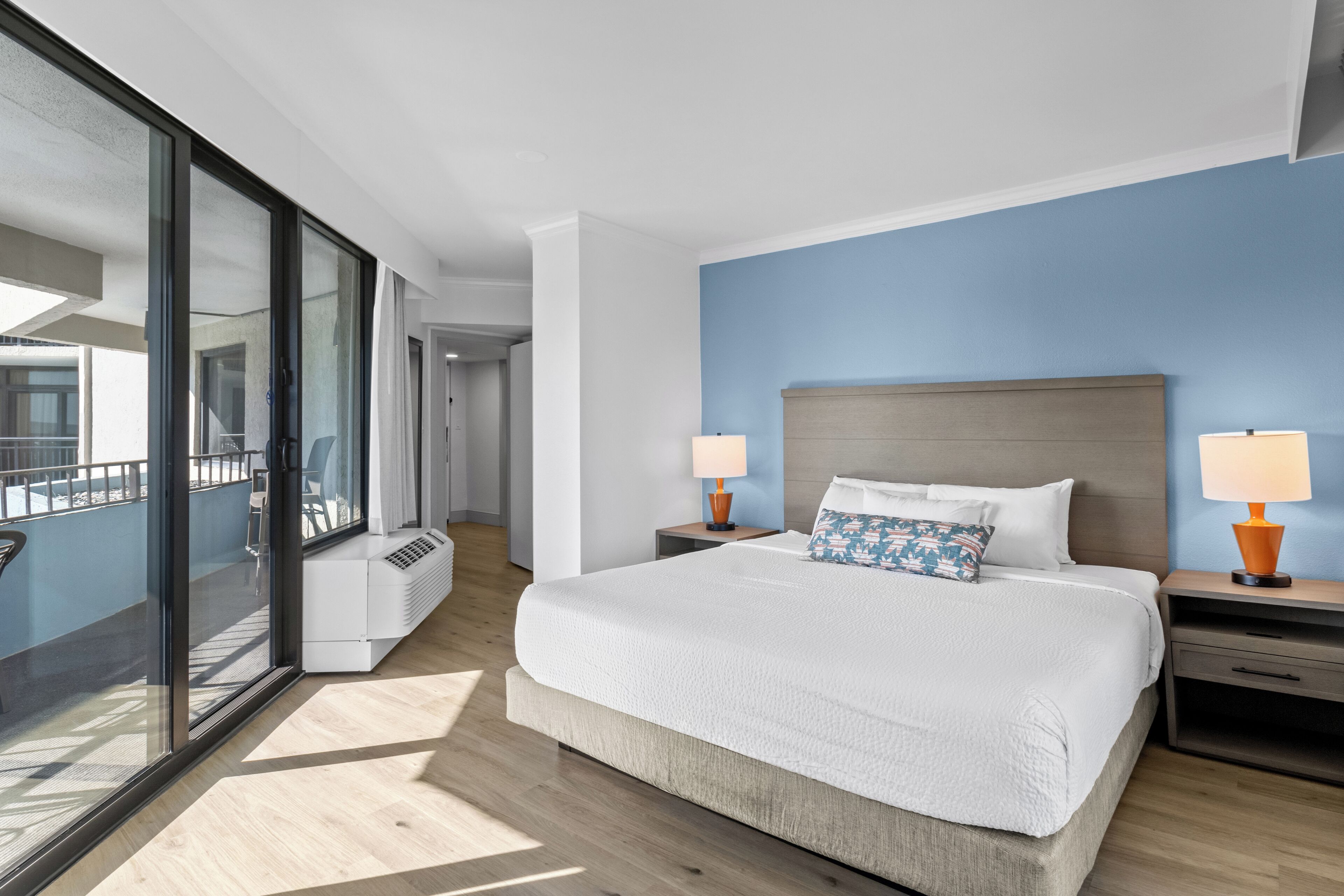 A modern hotel room with a large bed, two bedside tables with lamps, a view through a sliding glass door to an outdoor balcony, and a blue accent wall.