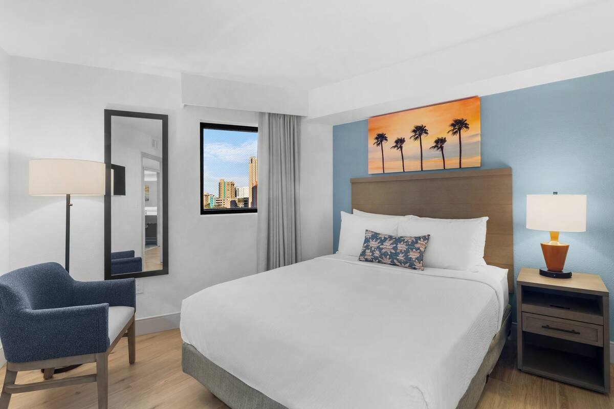 Quarto de hotel com cama, cadeira, mesa de cabeceira, luminária, pintura de palmeiras e janela com vista para a cidade.