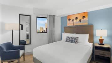 A hotel room with a bed, a chair, a nightstand, a lamp, a painting of palm trees, and a window with a city view.