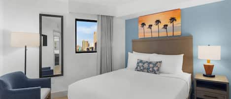 Hotelkamer met een bed, een stoel, een nachtkastje, een lamp, een schilderij van palmbomen en een raam met uitzicht op de stad.