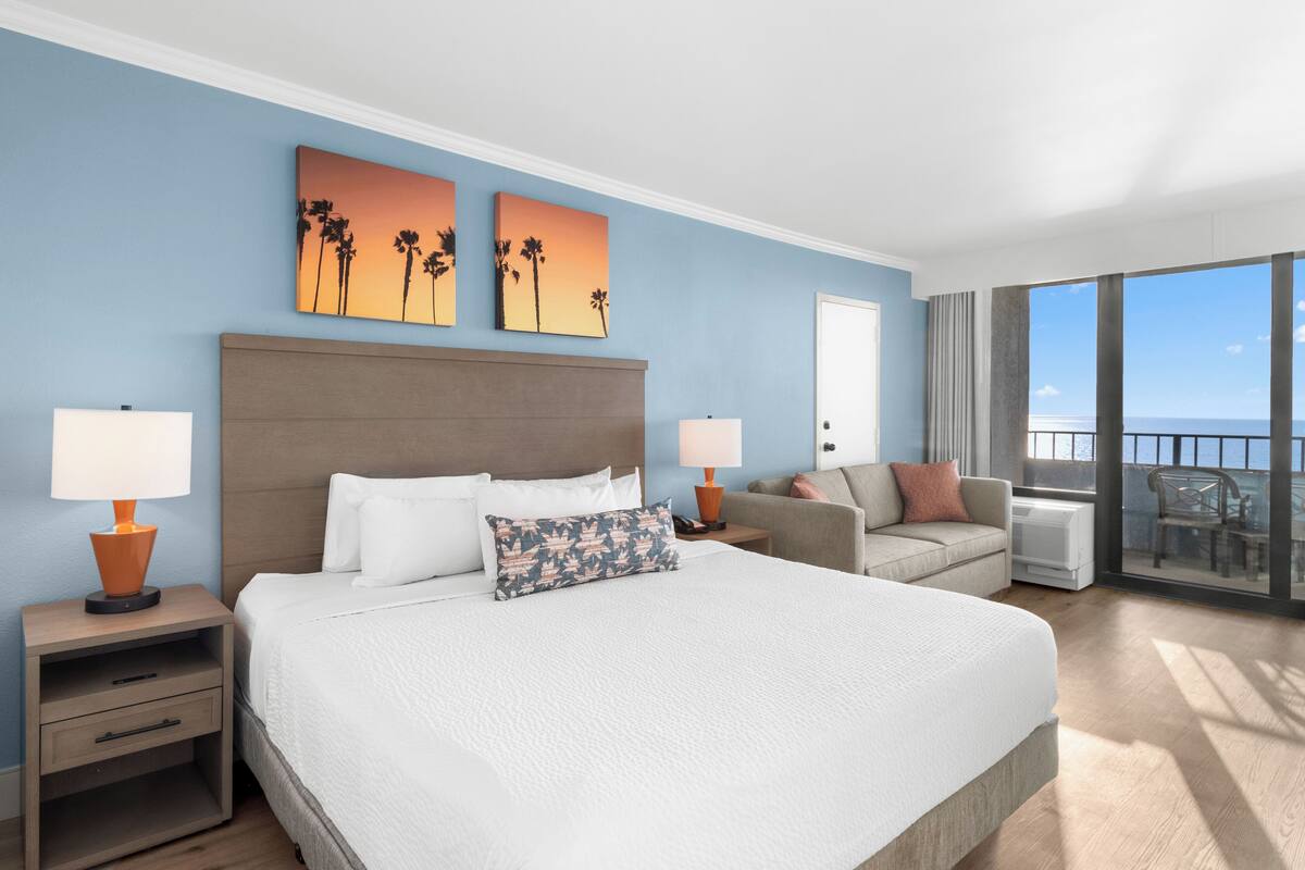 Quarto de hotel com uma cama grande, duas mesas de cabeceira, um sofá e uma varanda com vista.