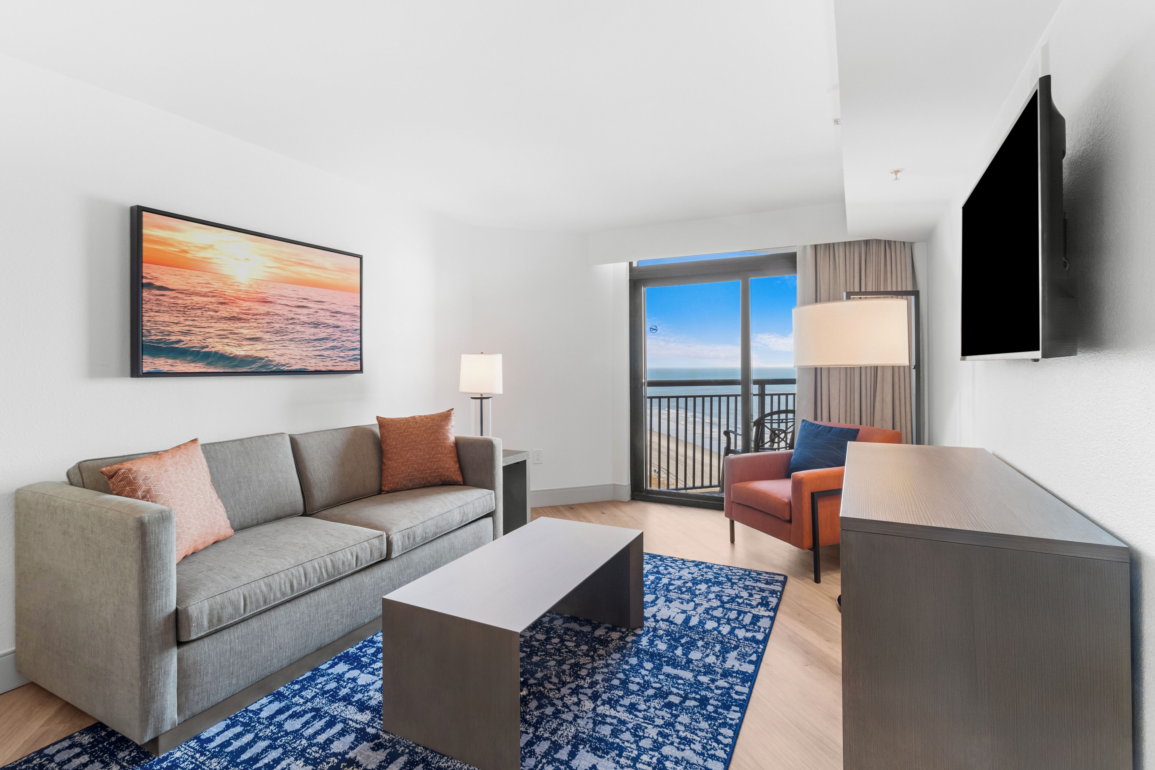 A modern living room with a grey sofa, a coffee table, and a blue patterned rug. There is a TV mounted on the wall and a window with a view of the ocean.