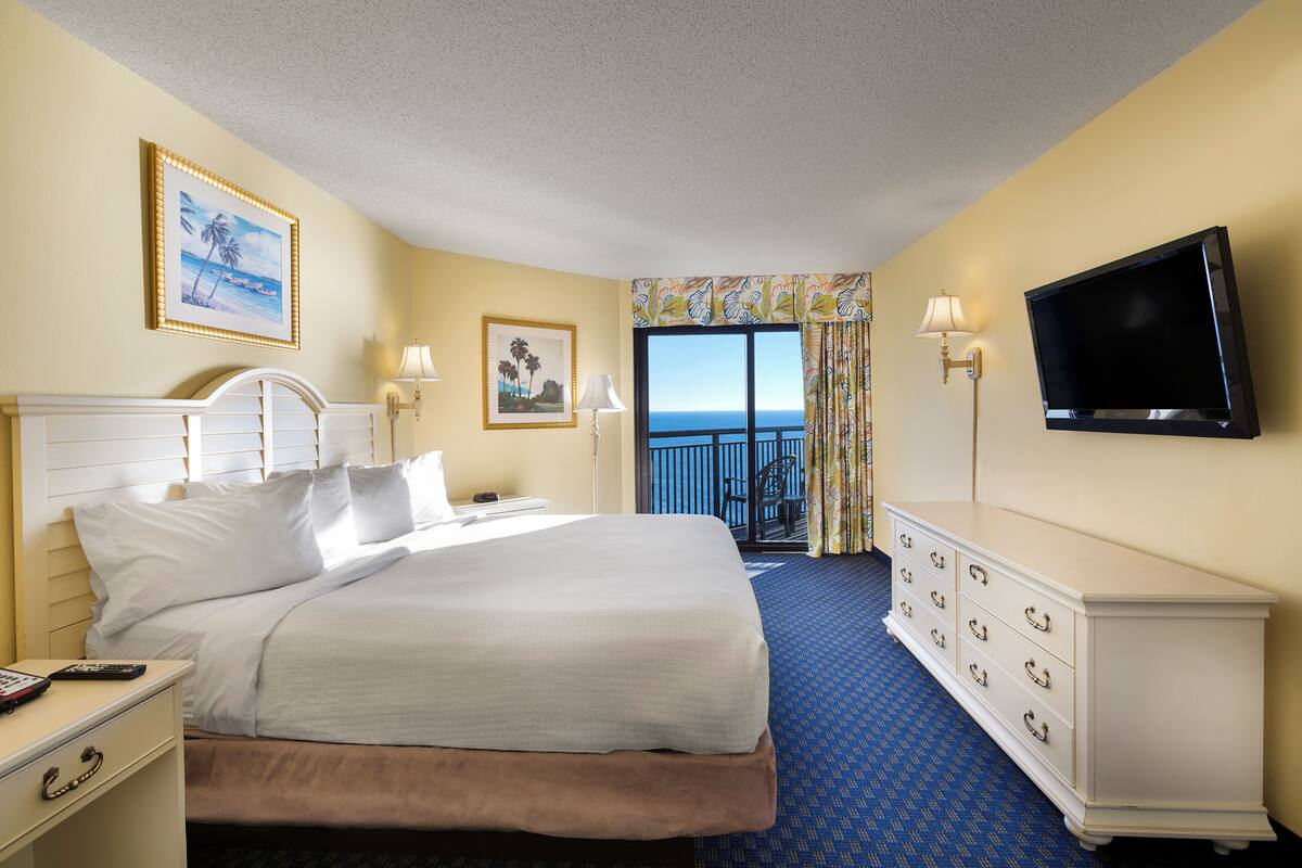 A hotel room with a bed, a dresser, a TV, and a balcony with a view.