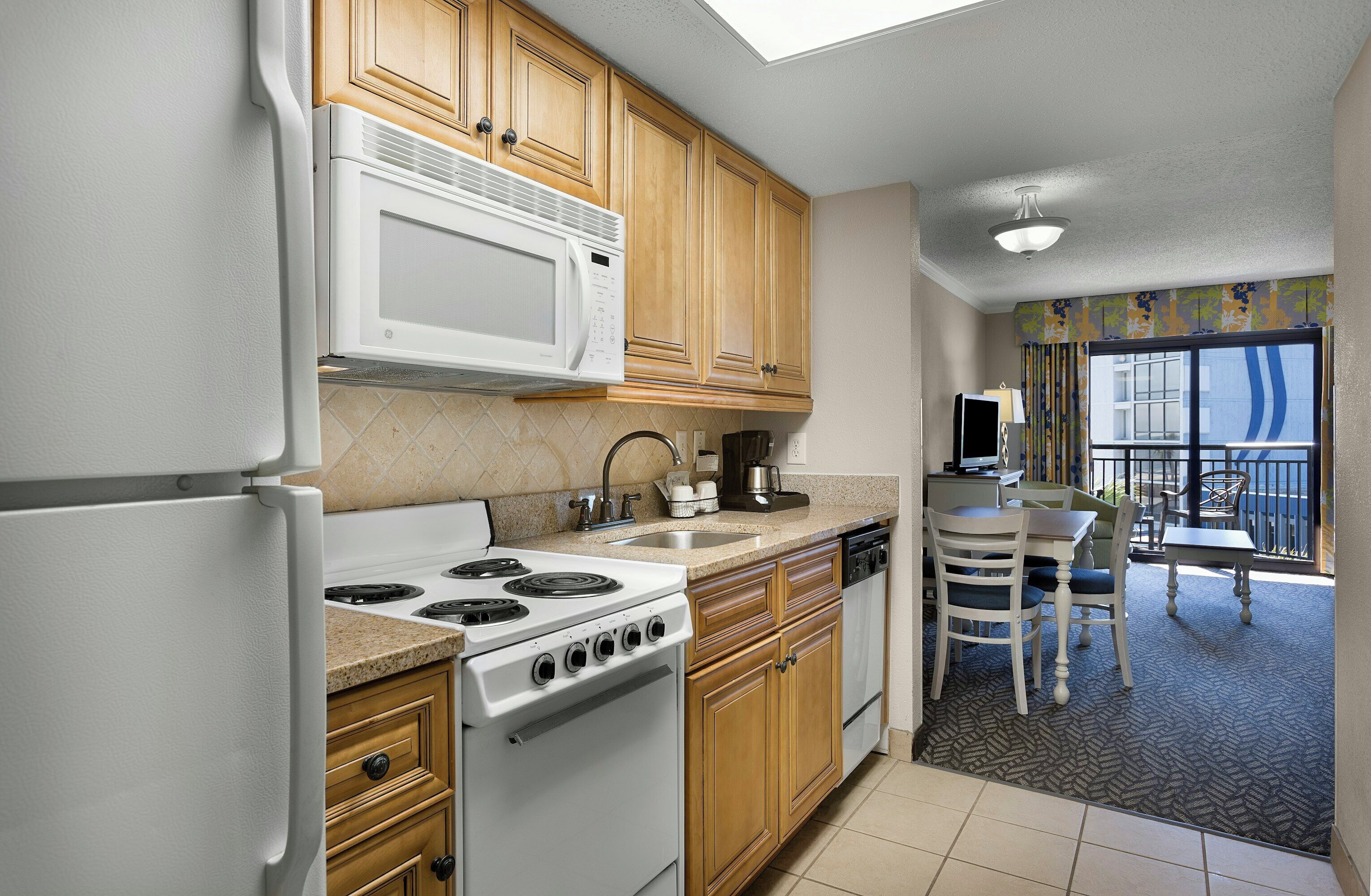 A compact kitchen with a stove, microwave, sink, and wooden cabinets. There is a dining area with a table and chairs, and a balcony with a view.