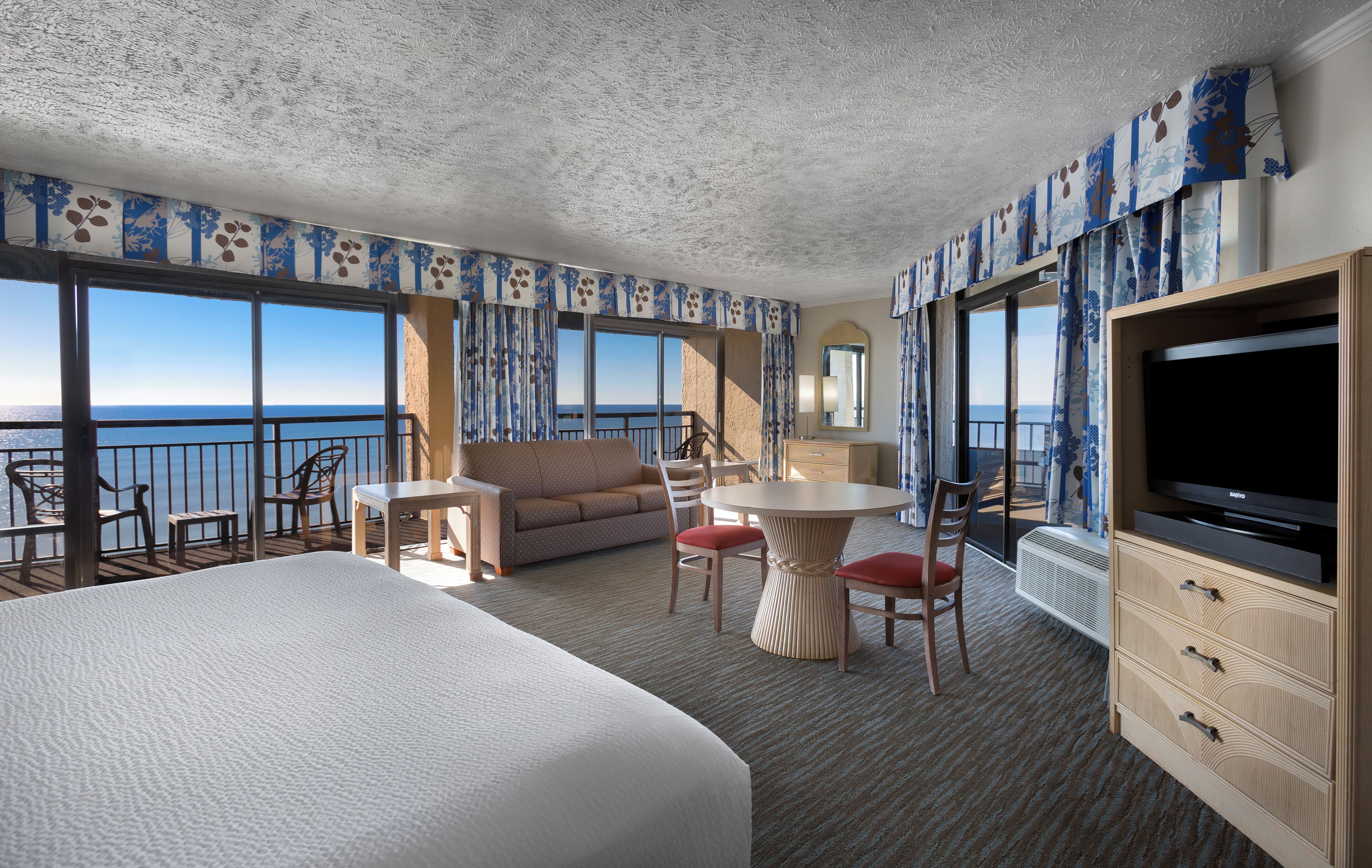 A hotel room with a large bed, a sofa, a dining table with chairs, a TV, and a view of the sea.