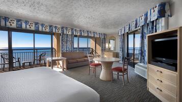 A hotel room with a large bed, a sofa, a dining table with chairs, a TV, and a view of the sea.