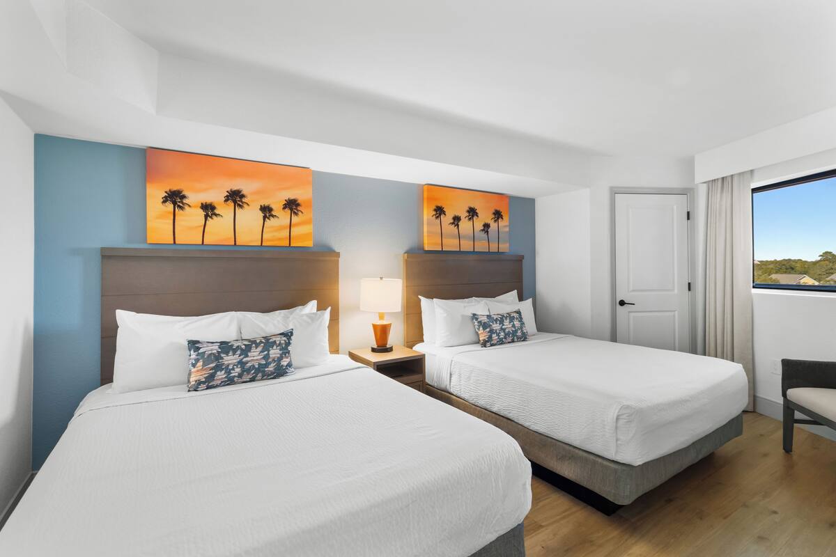 Quarto de hotel com duas camas, mesa de cabeceira com abajur e um quadro de palmeiras na parede.