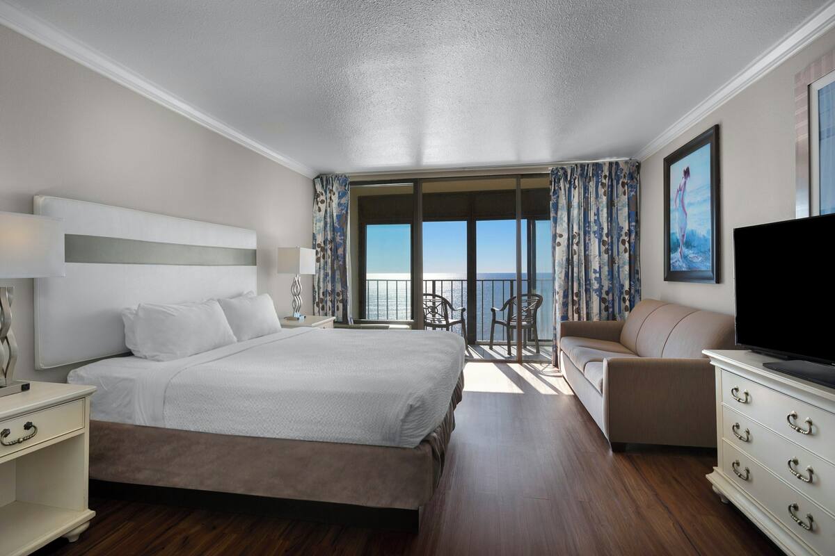 Quarto de hotel com uma cama grande, um sofá, uma televisão e uma varanda com vista.