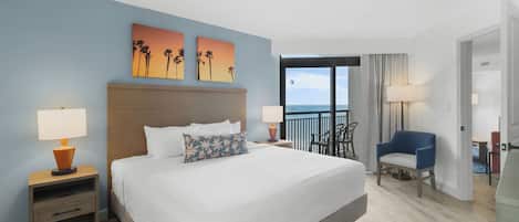 A hotel room with a large bed, two bedside tables with lamps, a view of the ocean, and a balcony with chairs.