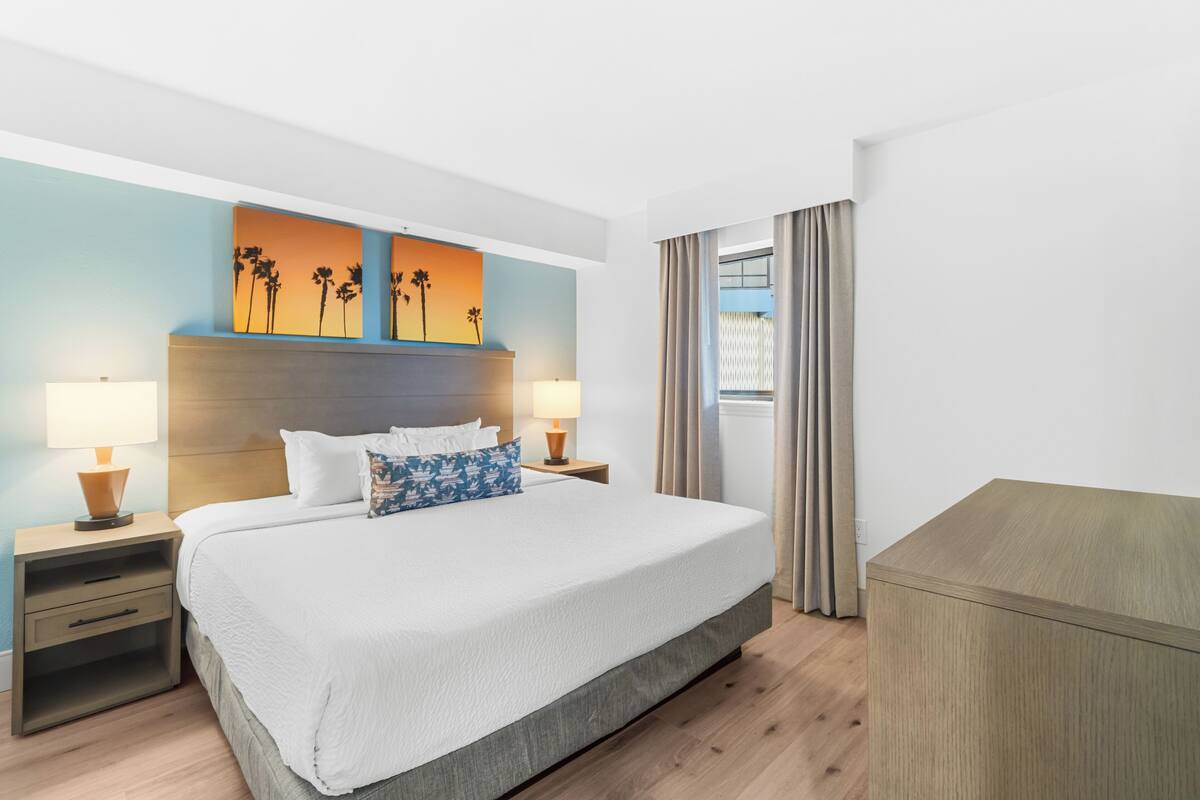 Quarto de hotel com uma cama grande, duas mesas de cabeceira com luminárias, um encosto de cama, vista para o exterior e uma cômoda de madeira.