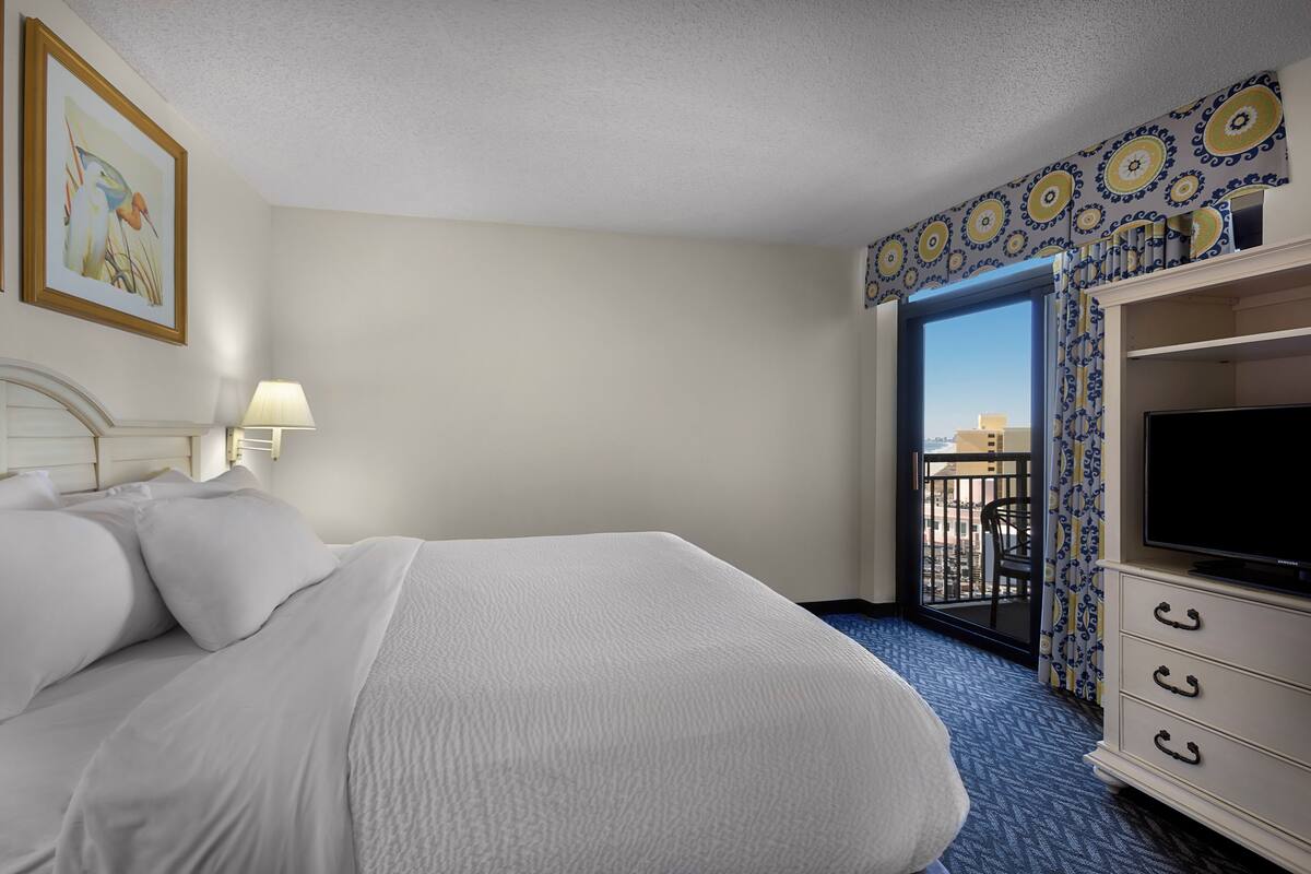 A hotel room with a bed, a TV, a balcony with a view, and a patterned wall.