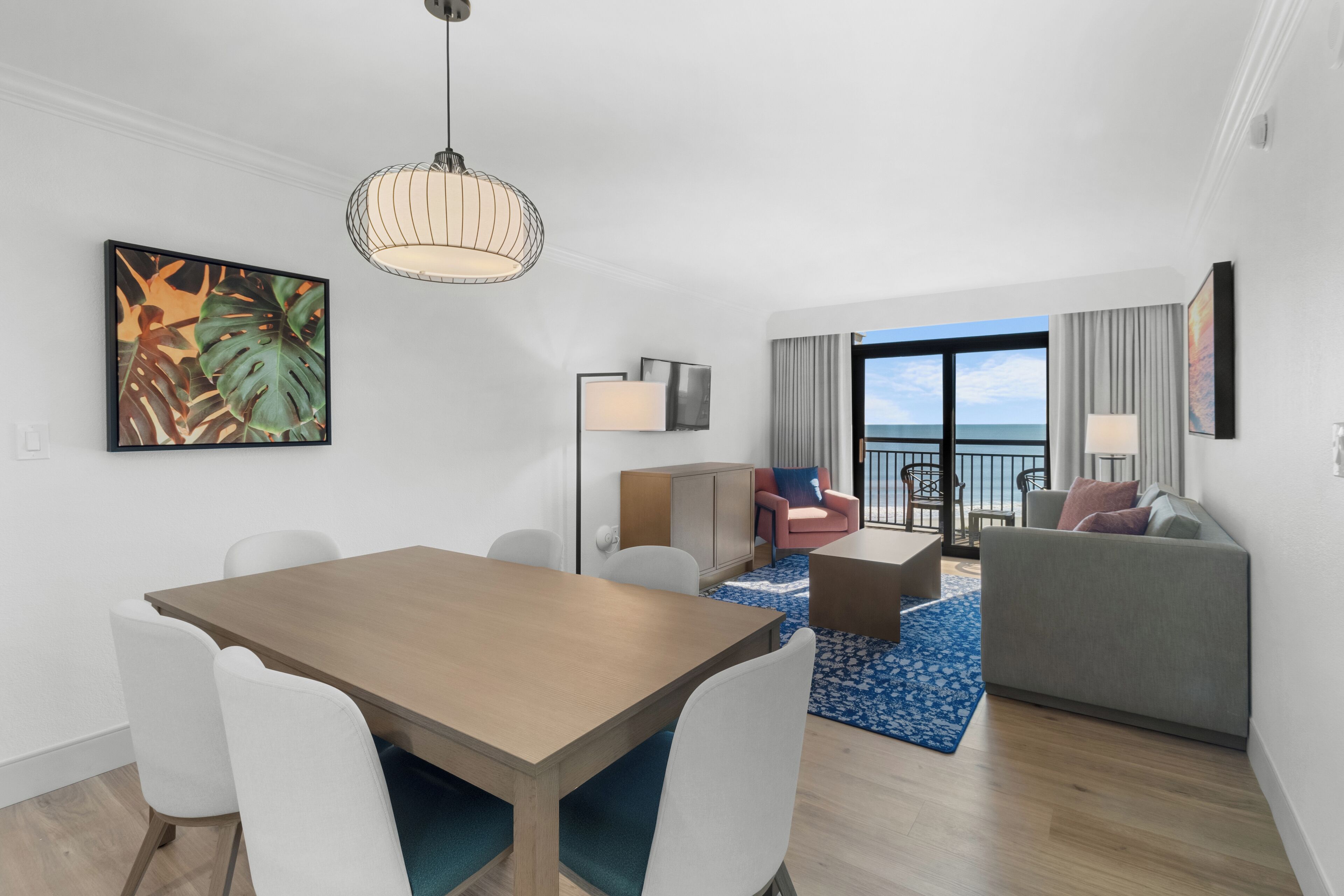 A modern hotel room with a dining area, a sofa, a balcony with a view, and a large window.