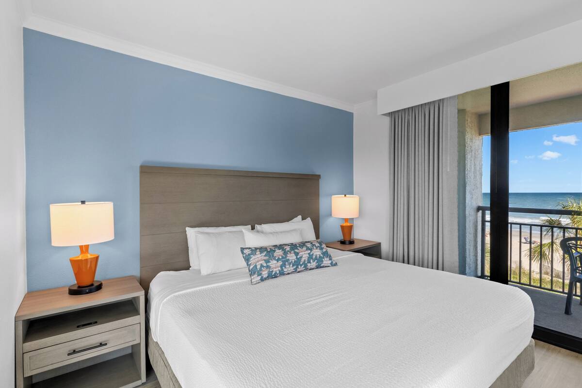Quarto de hotel com uma cama grande, duas mesas de cabeceira com luminárias, um criado-mudo com gaveta e vista para o oceano através de uma varanda.