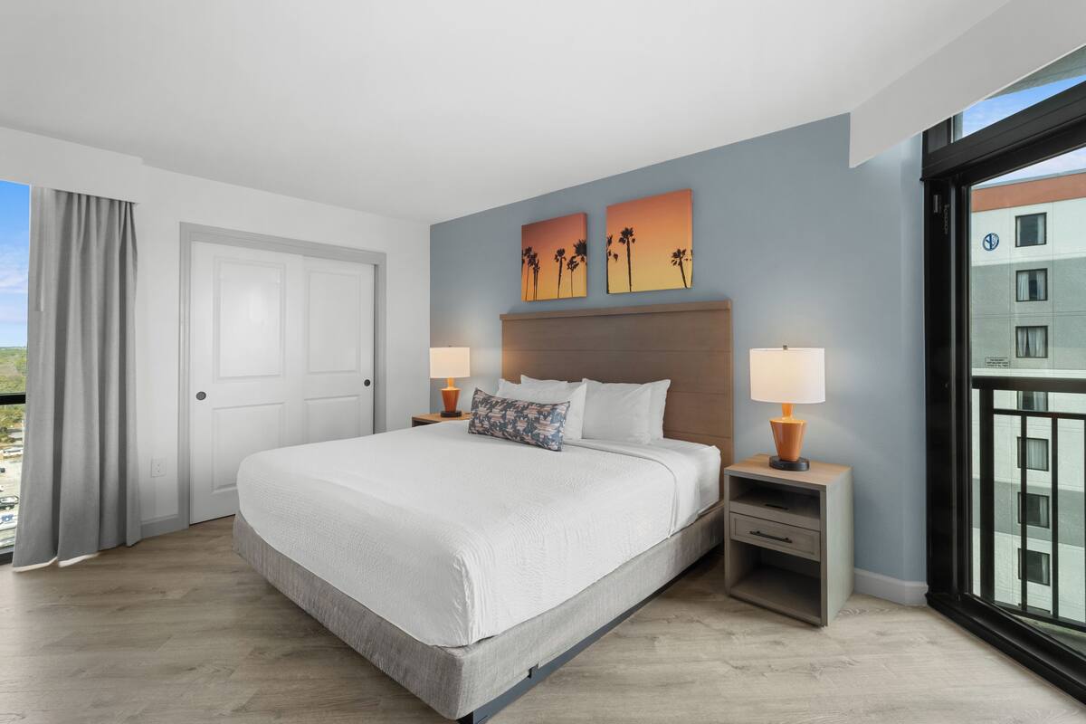 Quarto de hotel moderno com uma cama grande, duas mesas de cabeceira com luminárias, vista da cidade através de uma janela ampla e obras de arte na parede.