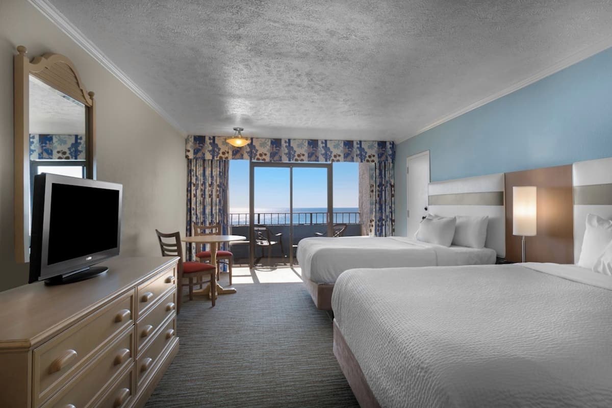Quarto de hotel com uma cama grande, televisão, escrivaninha com cadeira, mesa pequena e varanda com vista para o mar.
