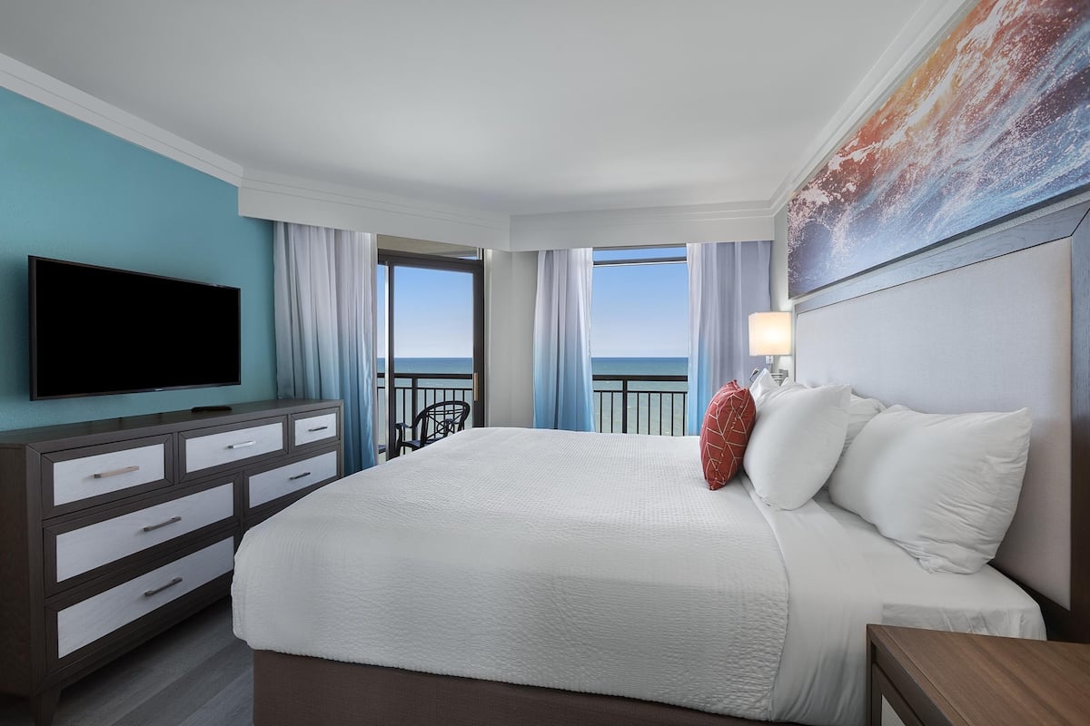 A hotel room with a large bed, a flat-screen TV, a dresser, and a view of the ocean through the windows.