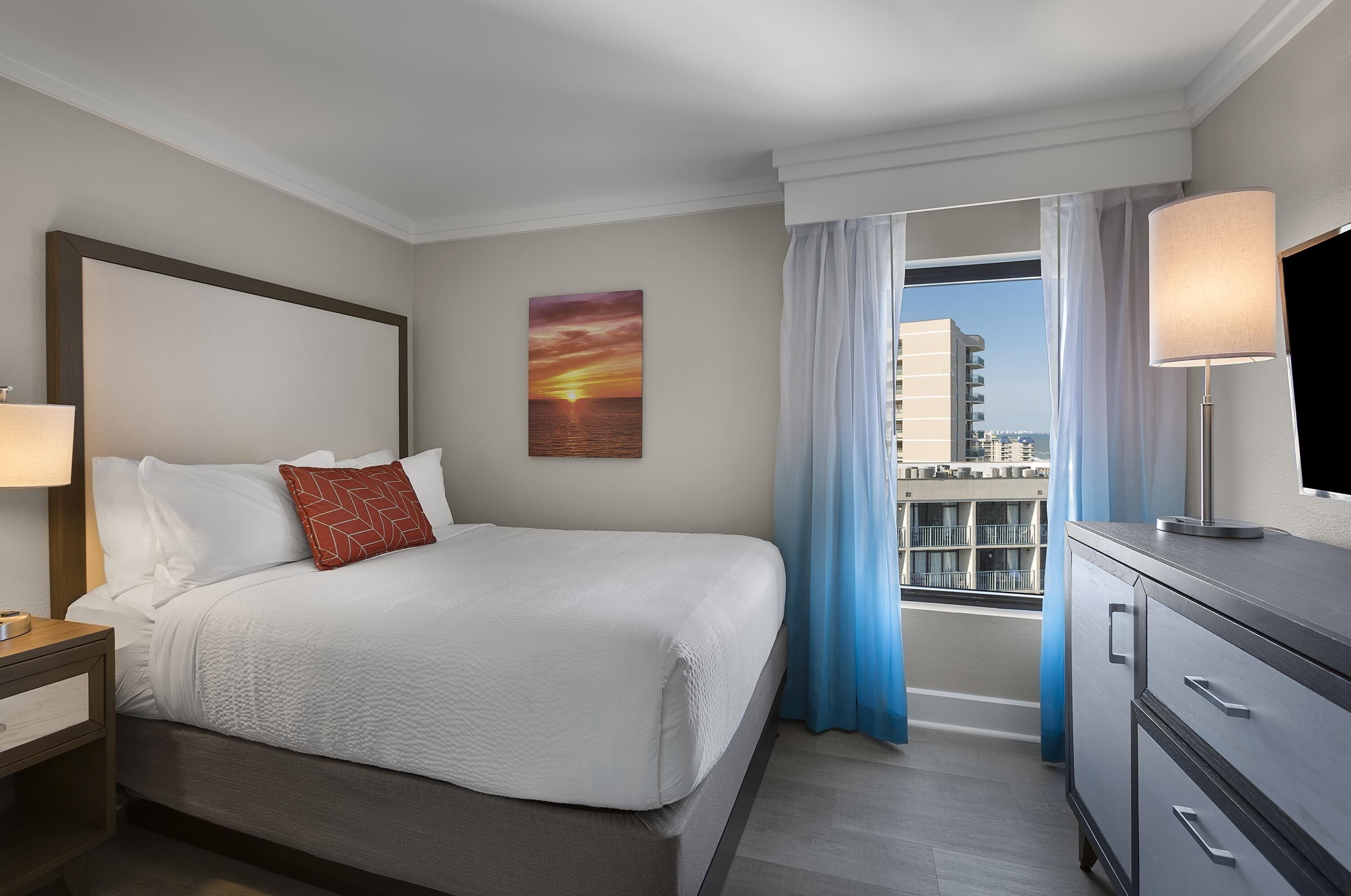 A hotel room with a large bed, a nightstand with a lamp, a TV on a dresser, and a window with curtains.