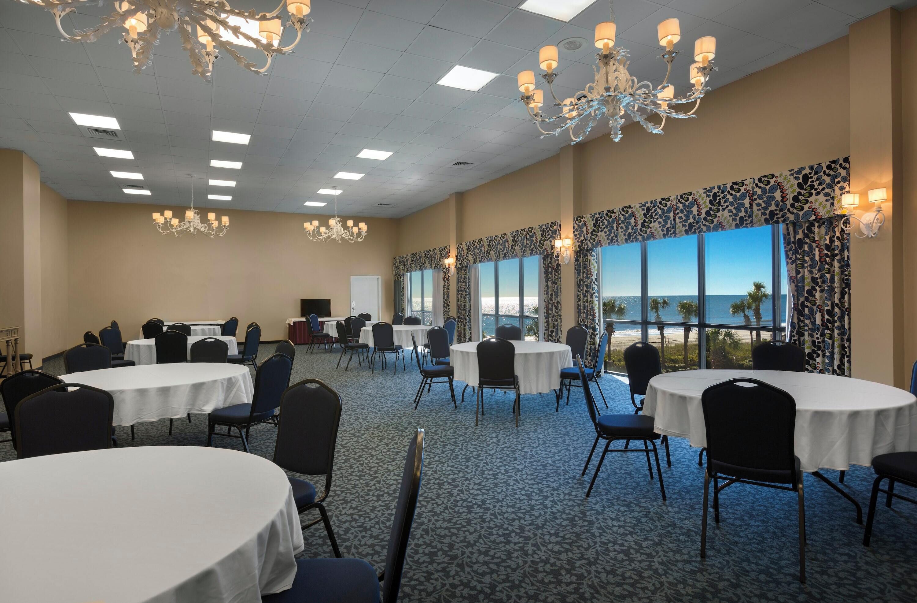 A conference room with round tables and chairs, large windows with a view of the beach, and chandeliers.