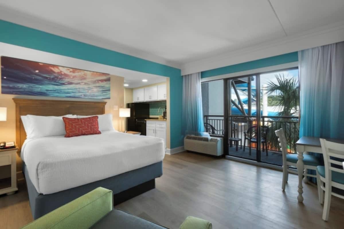 A hotel room with a bed, a desk, a balcony with a view of a pool, and a kitchen area.