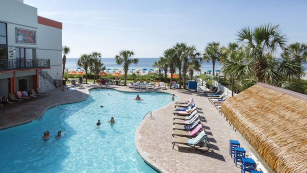 Piscina con sillas reclinables y sombrillas, una playa al fondo y un edificio con un letrero.