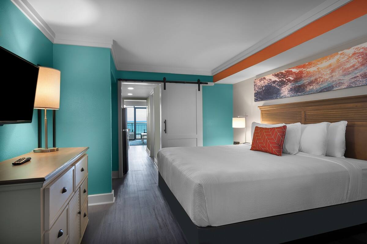 A hotel room with a bed, a TV, a desk, and a door leading to another room.