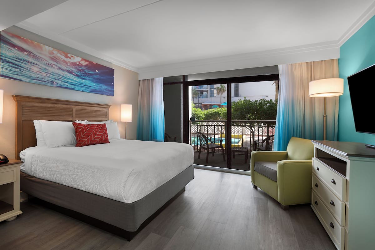 A hotel room with a large bed, a desk, a chair, a TV, and a balcony with a pool view.