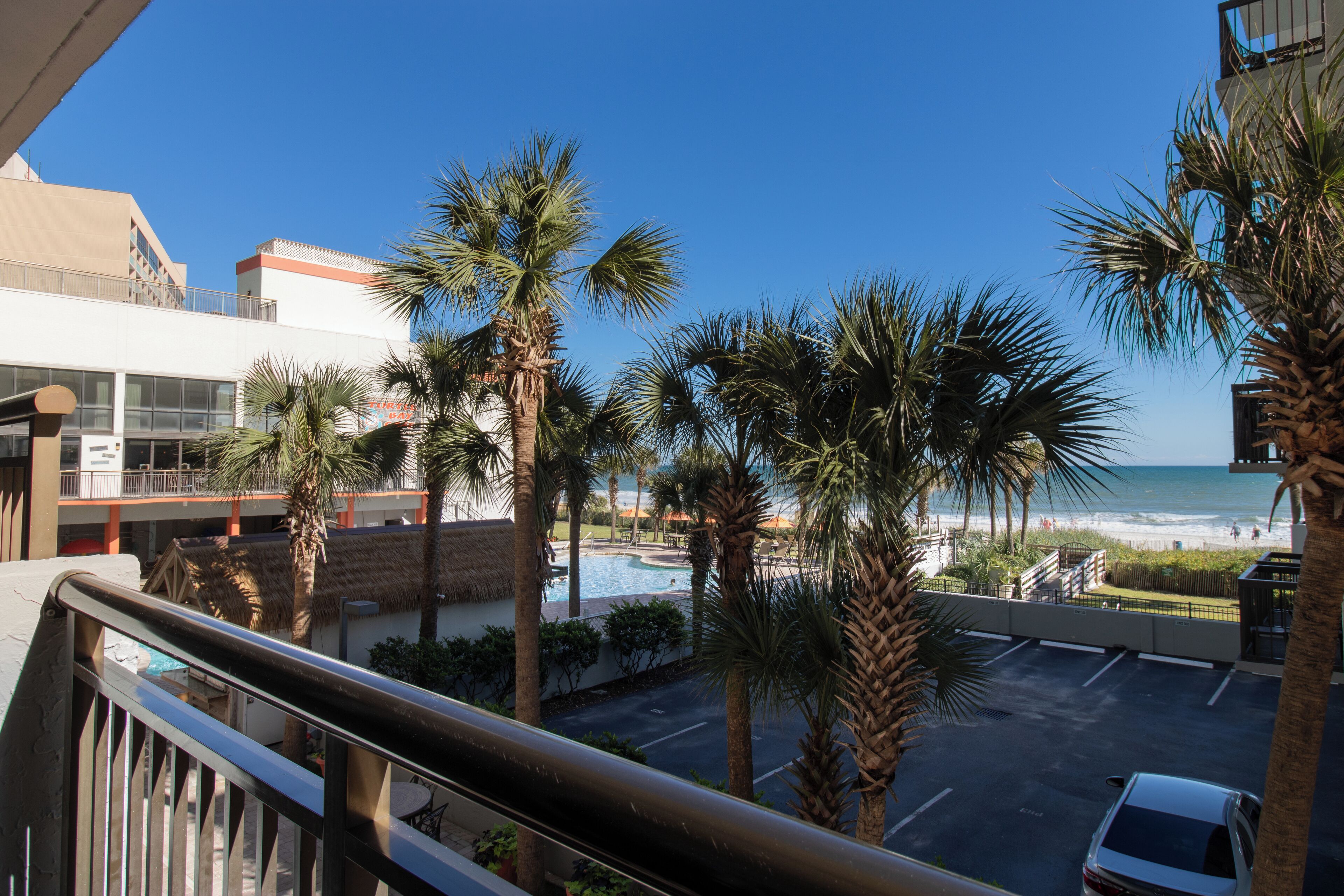 Blick vom Balkon auf einen Poolbereich mit Palmen, einen Parkplatz und einen Strand im Hintergrund.
