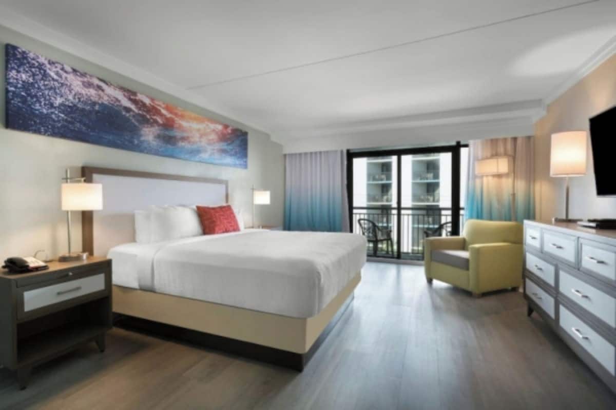A modern hotel room with a large bed, a desk with a lamp, a chair, a television, and a balcony with a view.