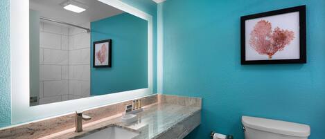 Un baño con un espejo grande, un lavabo con encimera de mármol, un inodoro y una obra de arte enmarcada en la pared.