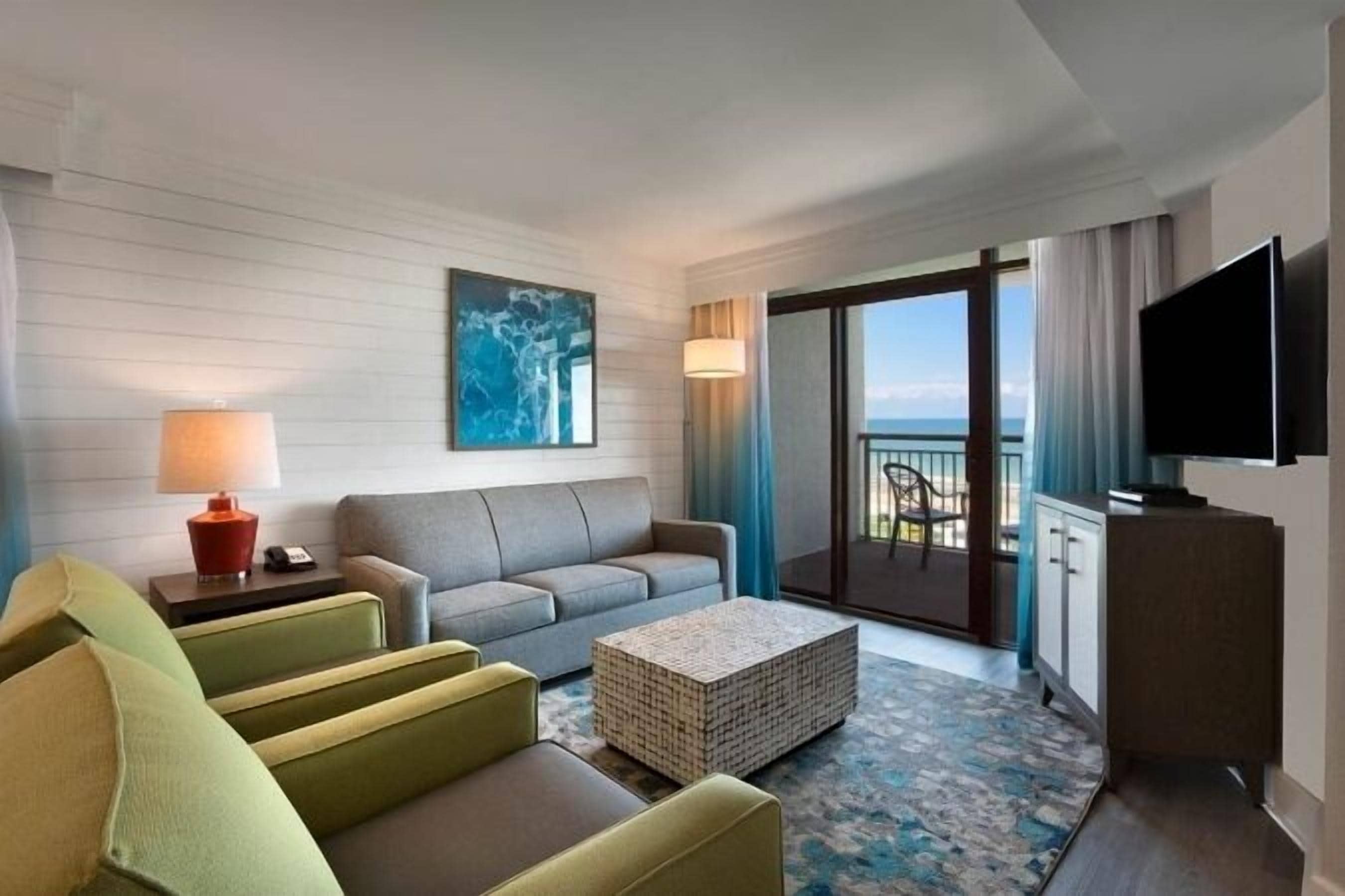 Habitación de hotel con sofá, mesa de centro, televisor de pantalla plana, balcón con vistas y un cuadro en la pared.