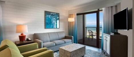Habitación de hotel con sofá, mesa de centro, televisor de pantalla plana, balcón con vistas y un cuadro en la pared.