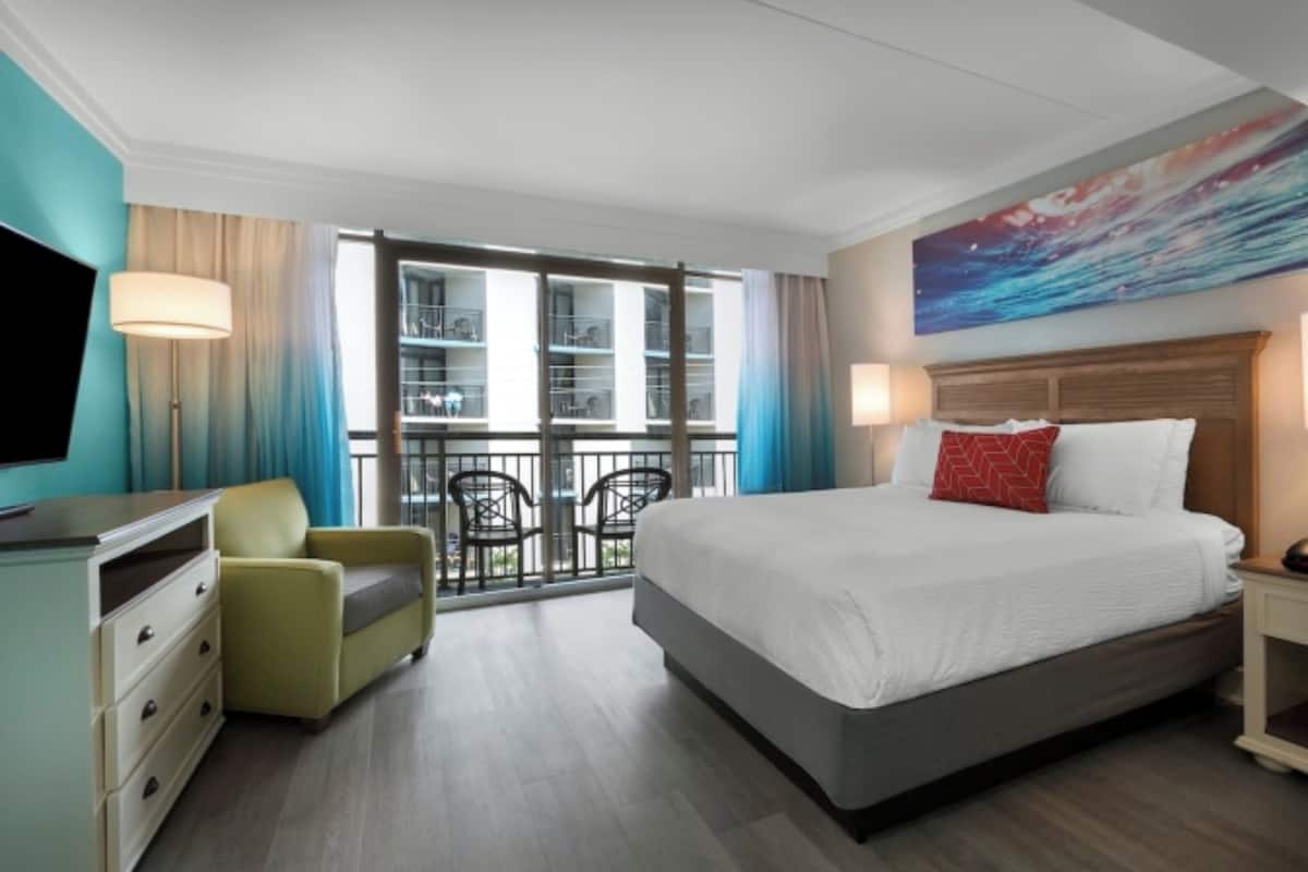 A hotel room with a bed, a TV, a chair, a desk, and a balcony with a view.