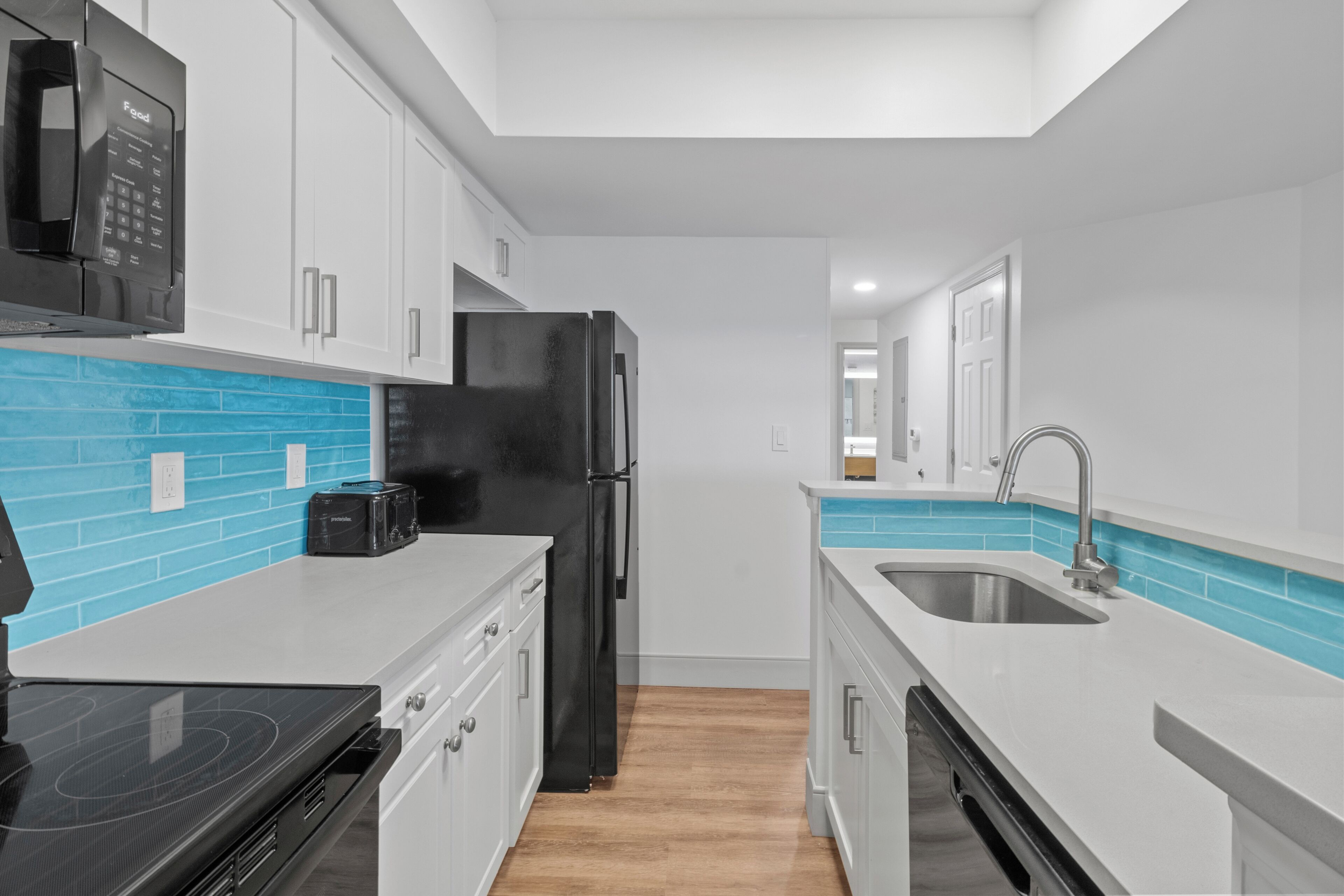 A modern kitchen with white cabinets, a black refrigerator, a microwave, a stove, and a sink with blue tiles.