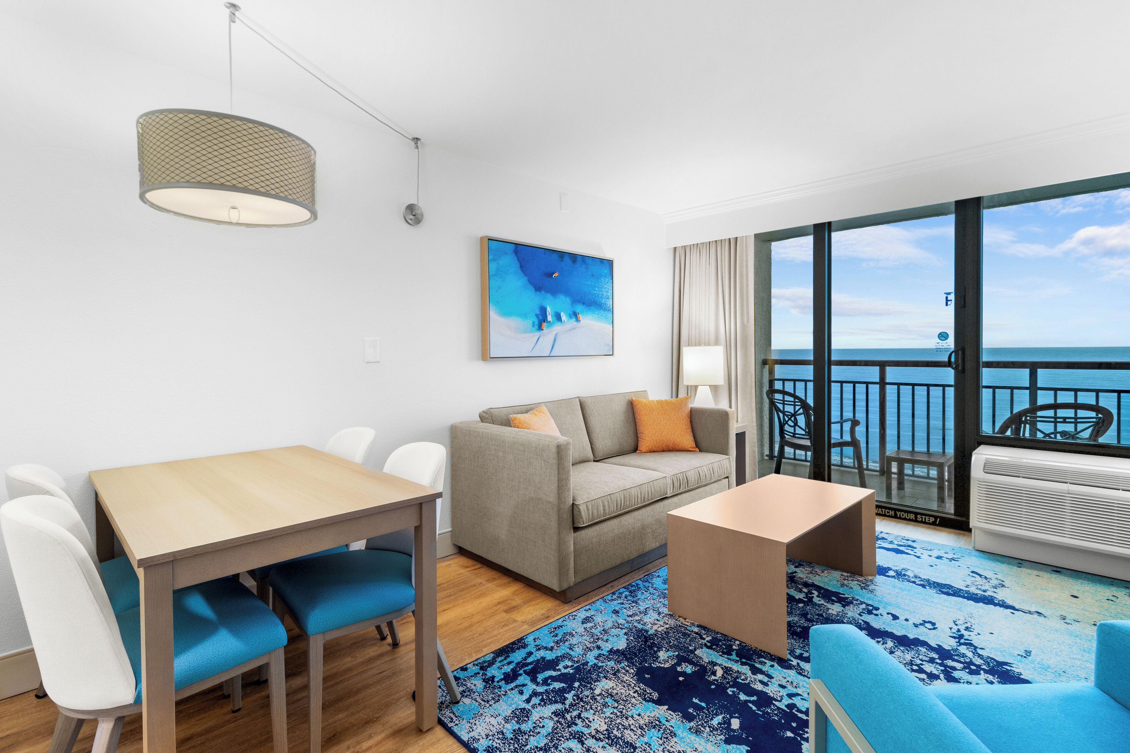 A modern living room with a sofa, coffee table, and dining table. There is a large window with a view of the ocean.