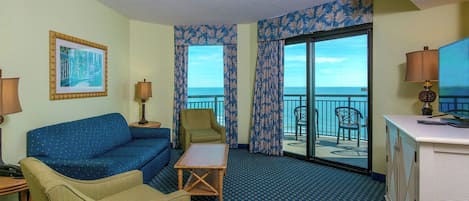 A hotel room with a blue patterned carpet, a blue sofa, a wooden coffee table, a green armchair, a desk with a lamp, and a large window with a view of the ocean.