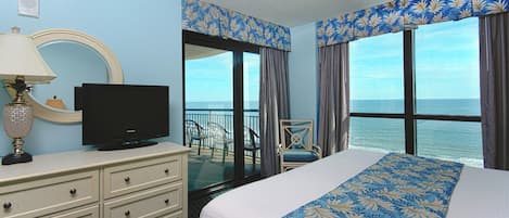 Una habitación de hotel con una cama, un televisor en una cómoda, una silla y vistas al océano a través de amplios ventanales.