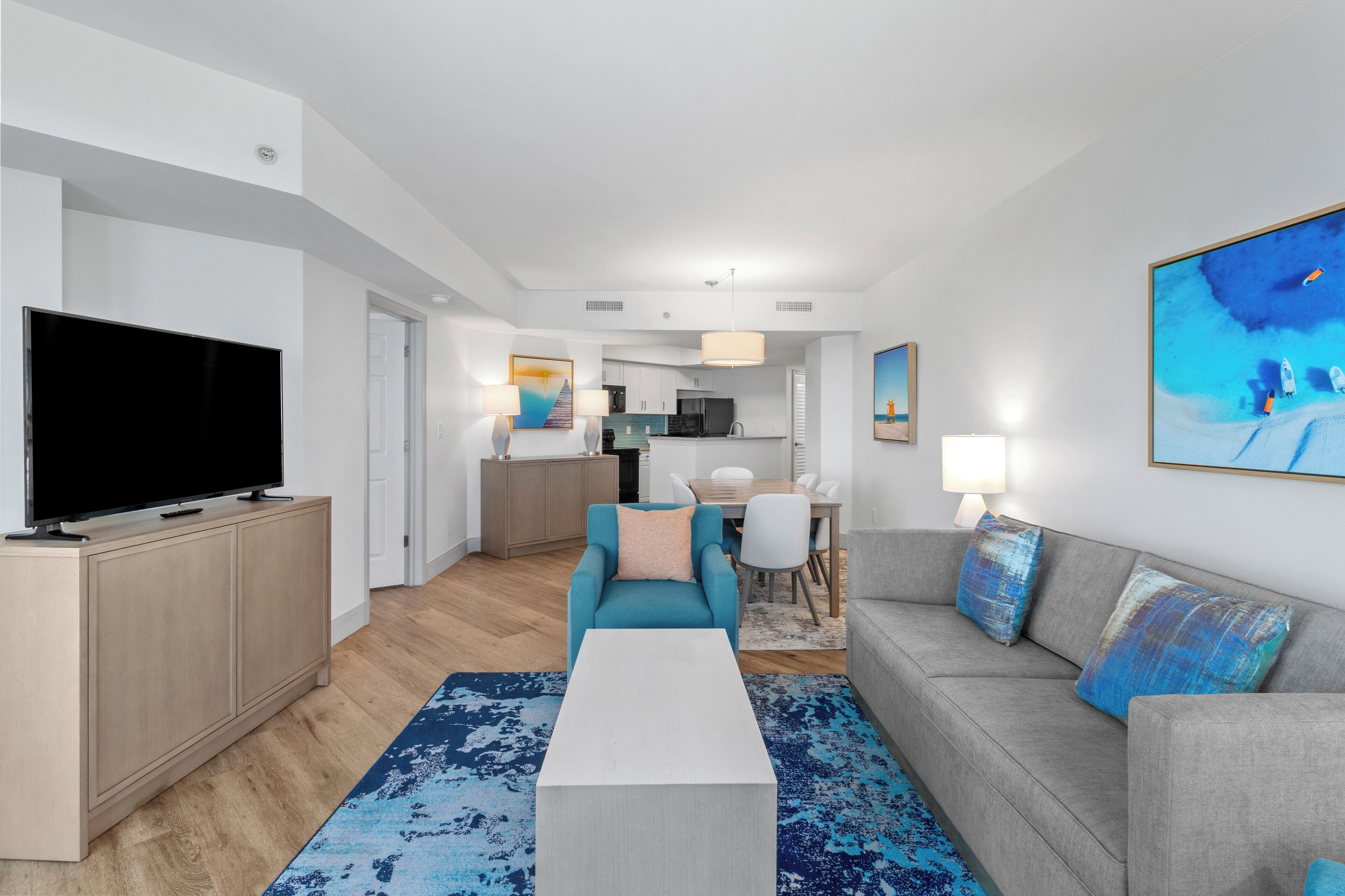 A modern living room with a flat-screen TV, a sofa, a coffee table, and a kitchen in the background.