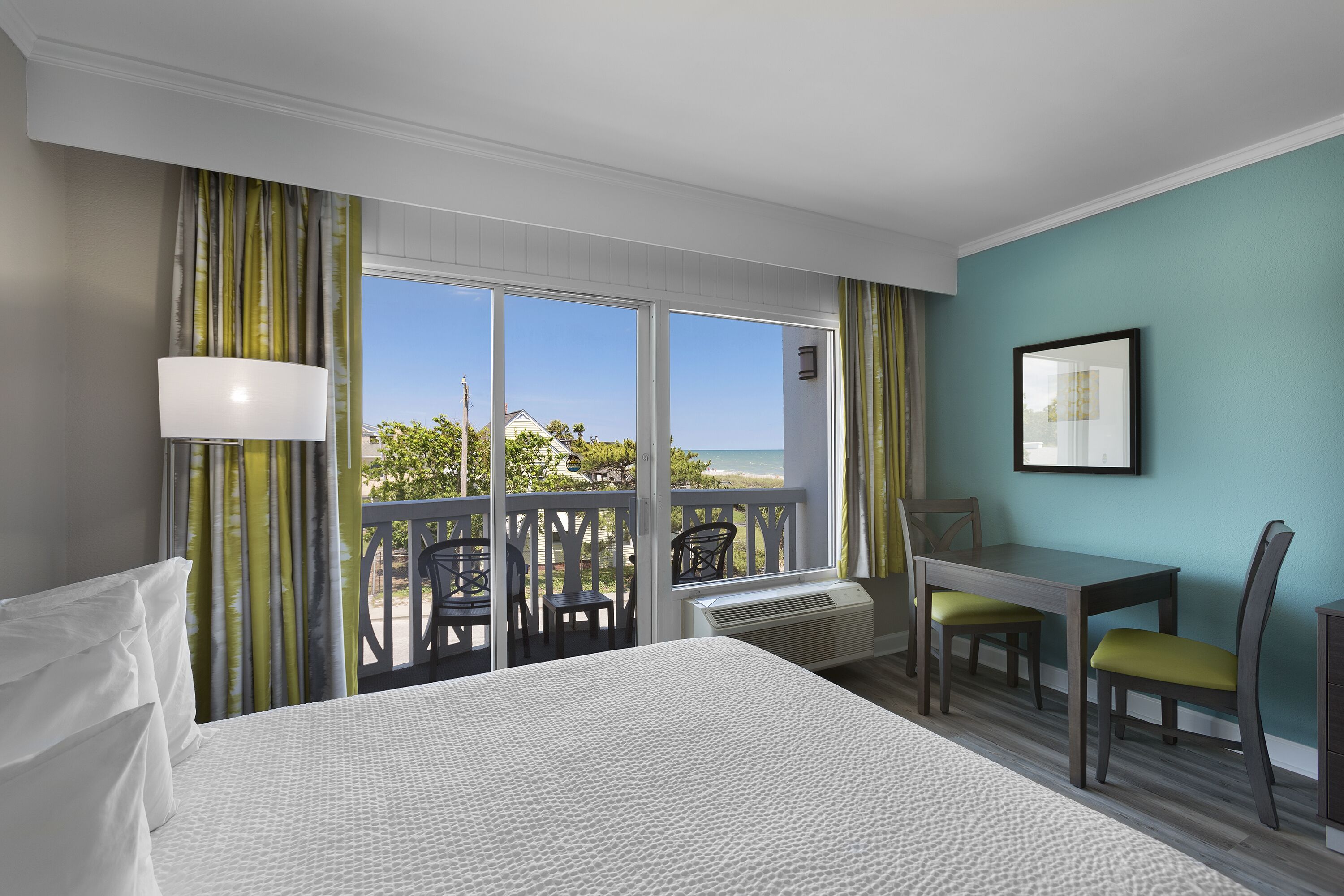 Une chambre d’hôtel comprenant un lit, un bureau, deux chaises, une fenêtre donnant sur l’extérieur et un tableau encadré au mur.