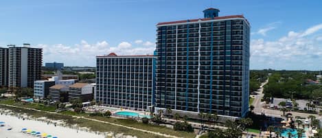 A high-rise hotel with multiple floors, a swimming pool, and a beach area with umbrellas.