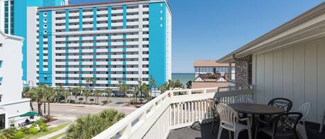 A balcony with a table and chairs, overlooking a road with palm trees and a large hotel building.