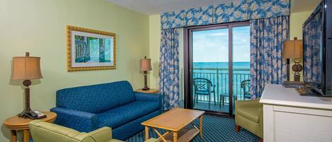 A hotel room with a blue sofa, a green armchair, a wooden coffee table, and a television.