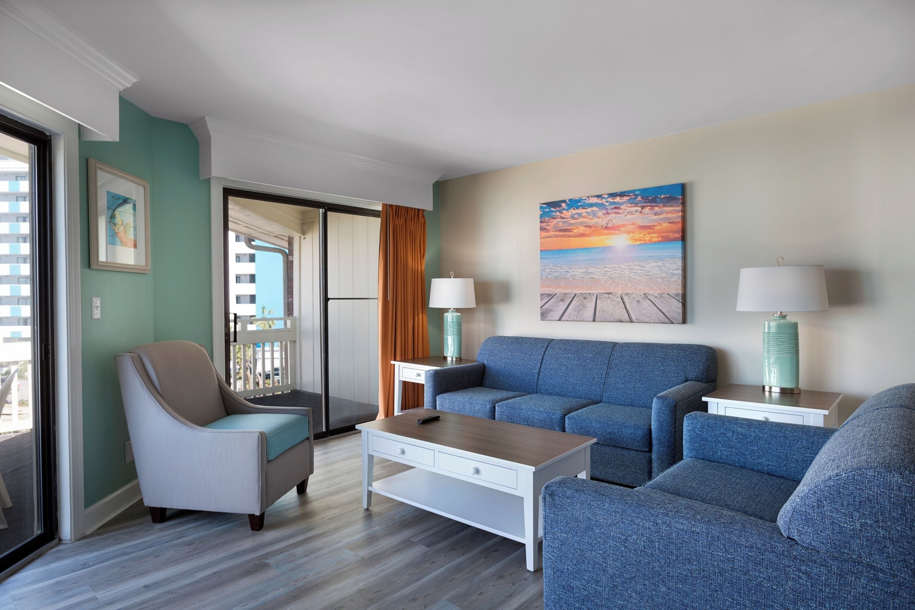 A modern living room with a blue sofa, a beige armchair, and a coffee table. There is a painting of a beach scene on the wall, and a window with curtains.