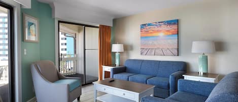 Un moderno salón con un sofá azul, un sillón color beige y una mesa de centro. En la pared, hay un cuadro de una playa, y una ventana con cortinas.