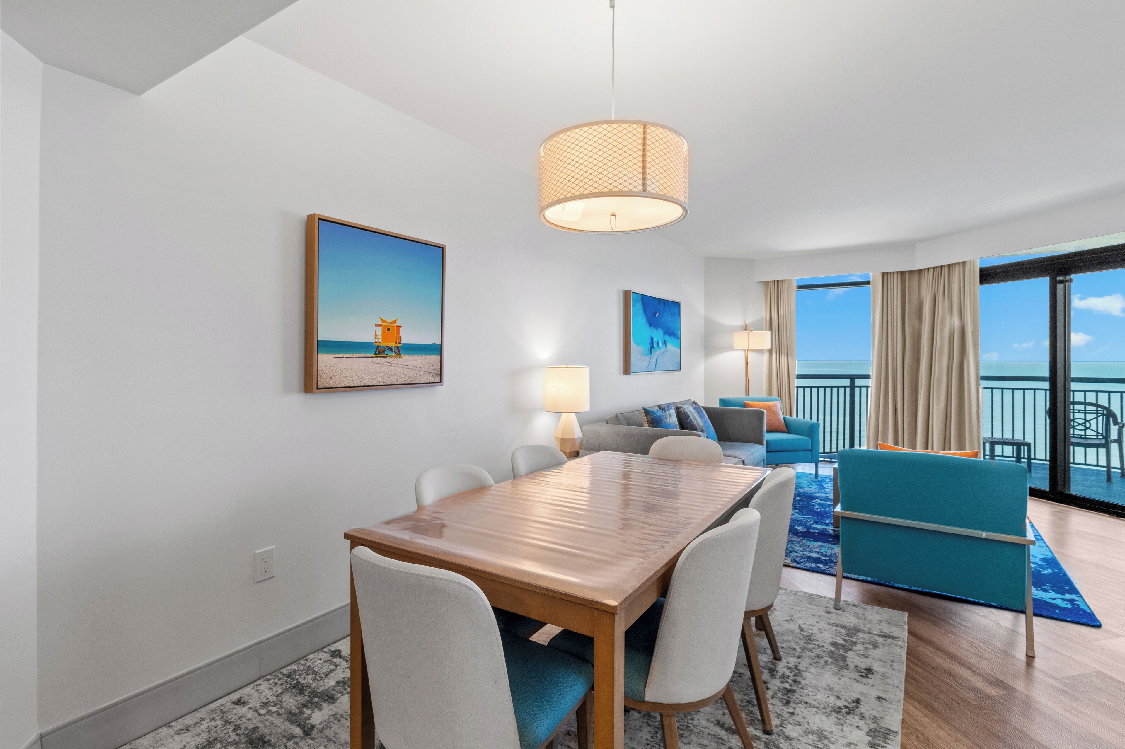A modern hotel room with a dining area, a sofa set, a balcony with a view of the sea, and artwork on the walls.
