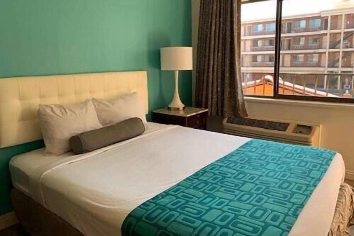 A hotel room with a bed, a nightstand, a lamp, a window with curtains, and a view of a building.