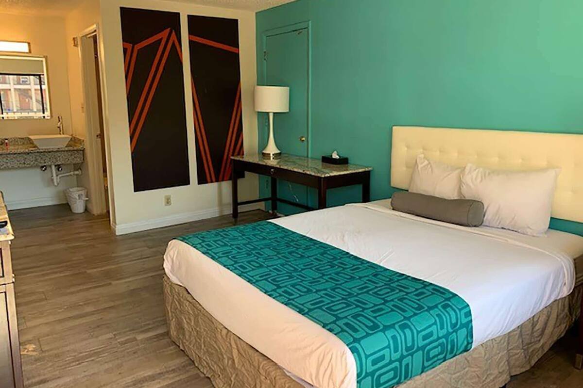 A hotel room with a bed, a nightstand, a lamp, and a bathroom visible in the background.