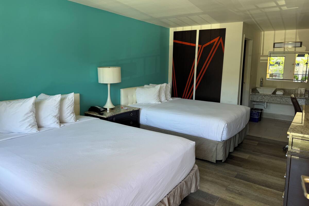 A hotel room with two beds, a desk with a lamp, a bathroom with a sink and mirror, and a teal wall.
