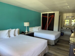 A hotel room with two beds, a desk with a lamp, a bathroom with a sink and mirror, and a teal wall. - Howard Johnson by Wyndham Reno Downtown (Reno)